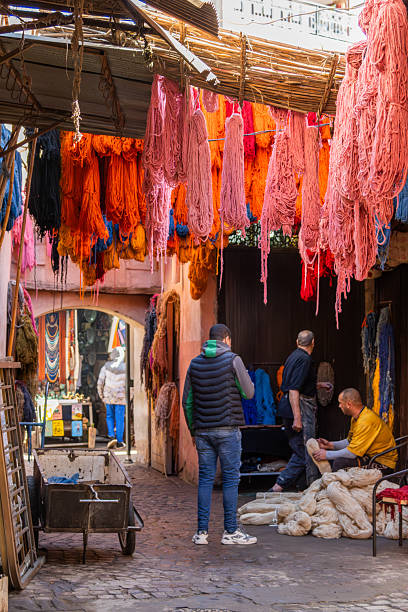 Vivid skeins of yarn in bright colors hang outside a workshop in Morocco, depicting traditional handcrafts and market ambiance, with locals working in vibrant surroundings.