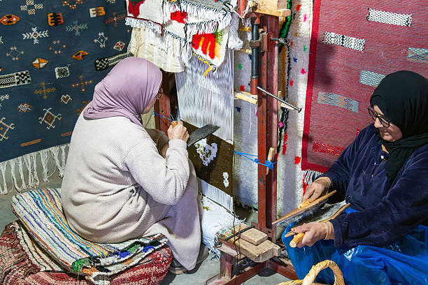 Two women work on creating traditional Moroccan rugs, spinning yarn and weaving intricate patterns. The setting features colorful textiles and a focus on cultural craftsmanship.