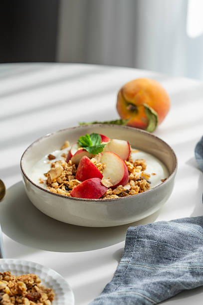 Natural greek yogurt with granola or muesli and peach in a bowl on a white table near window