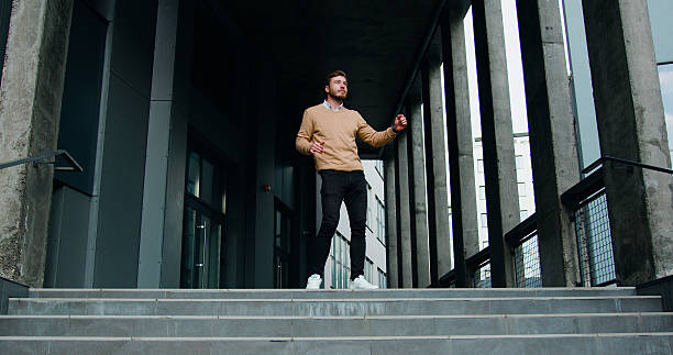 Happy and handsome caucasian young bearded businessman standing on the steps while leaving office center building and dancing as got new job of his dreams. Outside