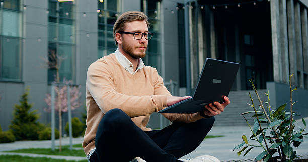istock Handsome young caucasian male working or studying using laptop while sitting crossing legs on bench of city financial district. Outdoors 2224470722