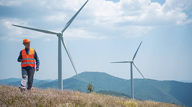 istock Power engineer wearing safety jacket and hard hat working at outdoor field site that have wind turbine at the background. 2224472937