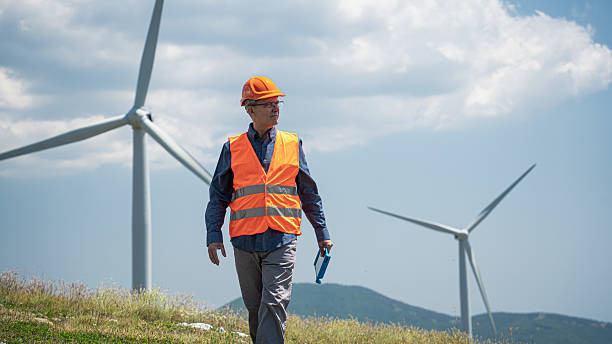 istock Power engineer wearing safety jacket and hard hat working at outdoor field site that have wind turbine at the background. 2224472964