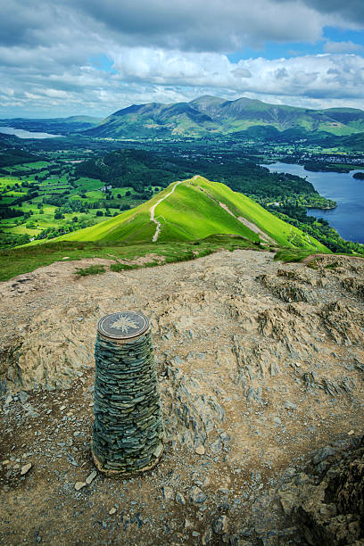 A vertical view looking down from the cairn at the summit of Catbells, where the ridgeline descends toward Derwentwater, surrounded by distant Lake District fells beneath a cloudy summer sky.