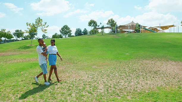 Parents walking with their child on a grassy hill under a clear blue sky near a playground