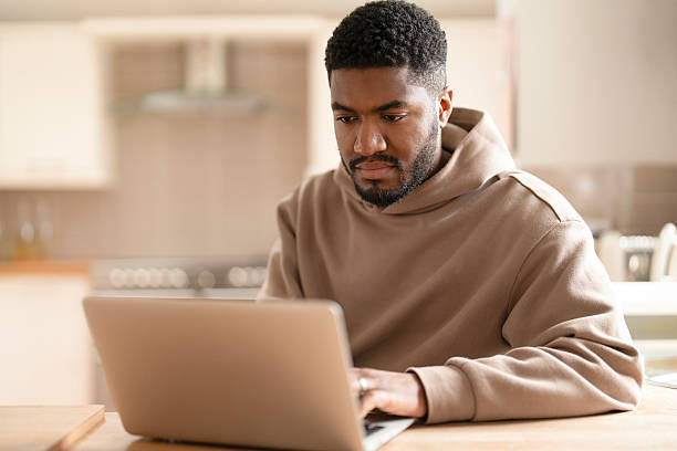Focused young man working on laptop at home in a cozy kitchen setting during the afternoon hours, engaged in online tasks or study