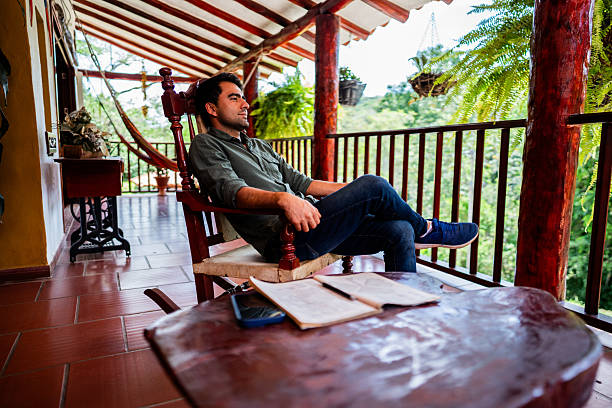 Young lonely teenager boy drinking hot tea from thermo flask while he sitting on the wooden forest house terrace and enjoying the morning spruce foggy forest. Traveling and tourism at fall time.