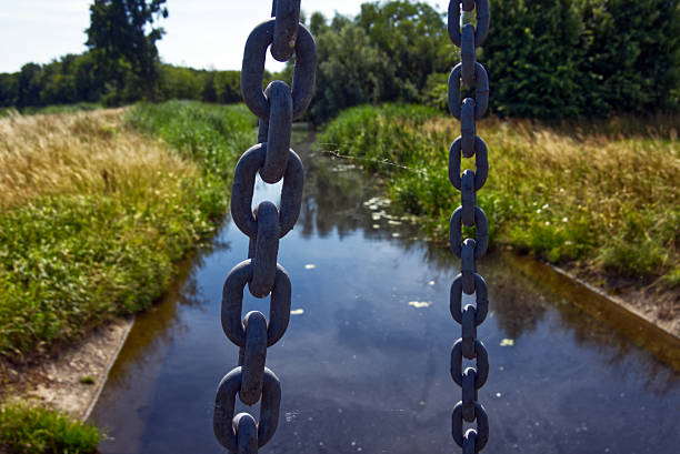 Two heavy, dark metal chains hanging above a drainage ditch create a striking contrast against the blurred greenery and water, suggesting a drawbridge mechanism