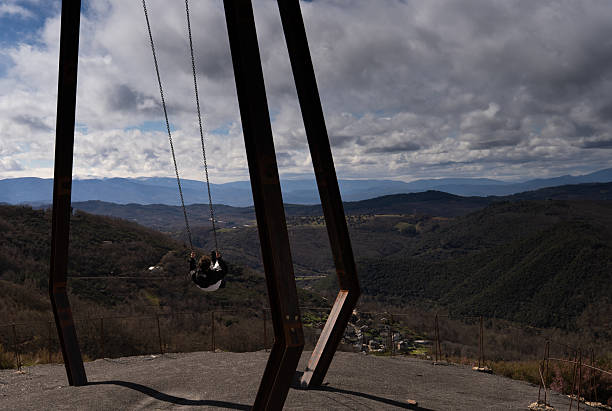 Teenage boy swinging from a large metal swing set overlooking the mountains and valleys of Libran, Leon, Spain. Captured under dramatic clouds, the image evokes freedom, elevation, and emotional connection with nature. Ideal for travel, lifestyle, and inspirational storytelling themes