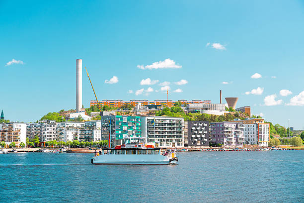 Ferry navigating the waters of stockholm, with colorful buildings and industrial structures in the background, on a bright summer day