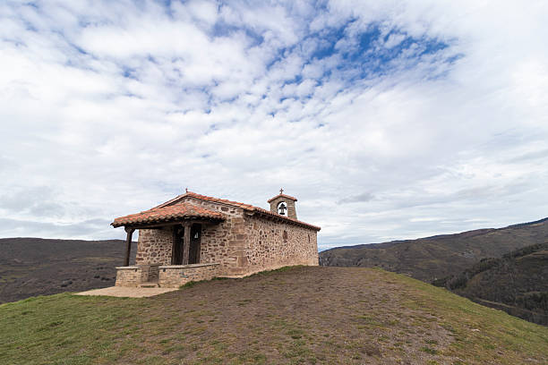 Small stone church with tile roof and bell tower standing on hill overlooking valley in spanish countryside