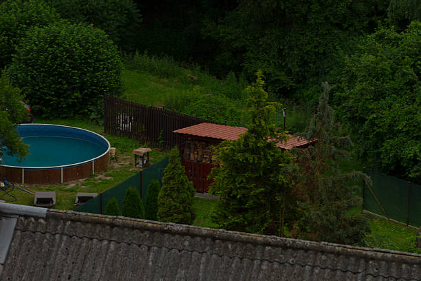 An aerial view shows a circular outdoor pool next to a charming wooden shed, surrounded by lush trees and soft grass, creating a serene setting for relaxation and leisure