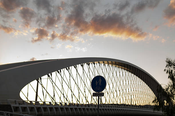 This image shows a scenic view of a historic bridge spanning a river in a city. The bridge has multiple arches and is reflected in the calm water below. The cityscape in the background includes various buildings, some with domes and classical architecture, indicating a European city, possibly Rome. The overcast sky adds a dramatic effect to the scene. The architectural beauty and historical significance of the location are beautifully captured, along with the serene atmosphere created by the reflection in the river.