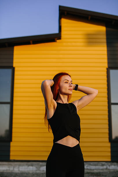Athletic woman with red hair stretching in front of a yellow and black modern building, enjoying outdoor exercise in the evening light
