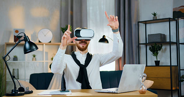 istock Handsome smiling young bearded man in virtual reality headset working on imaginary screen on the beautifully decorated home office background 2224517982