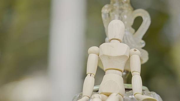 Poseable wooden mannequin figure seated outdoors with a soft, blurred green background.