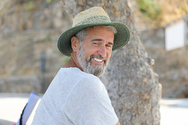 Smiling senior man with gray beard wearing a straw hat and white T-shirt, sitting on a bench under a tree on a sunny day. Cheerful expression and relaxed body language suggest a carefree summer vacation atmosphere.