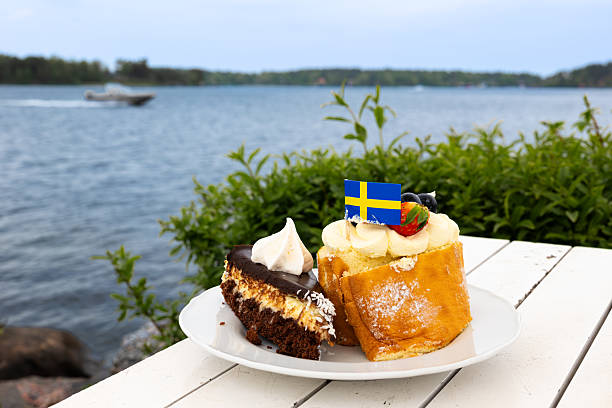 A plate with two types of cake, one chocolate and one cream, decorated with a Swedish flag, set against a scenic waterfront view at Vaxholm island, Sweden.