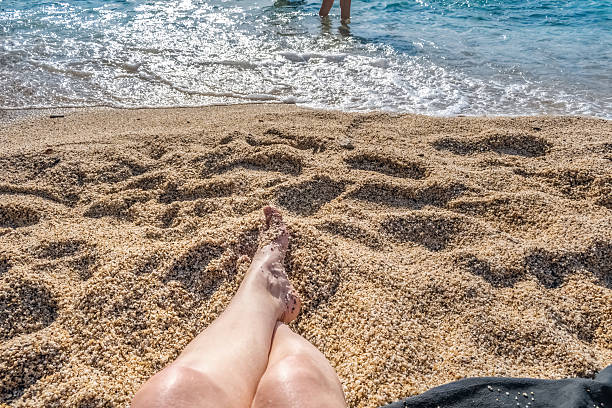 Relaxing on a sandy beach with feet resting in the sun as waves gently approach the shore. The scene captures a peaceful moment by the sea with sunlight sparkling on the water
