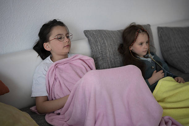 Two sisters snuggled under blankets on the couch watching TV. Concept: sibling bond, cozy evening, family time, shared moments.