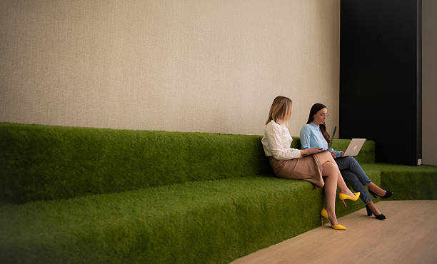 istock Two young businesswomen working on laptops while seated on modern green steps in a creative office. Focused and collaborative atmosphere with geometric wall art and natural textures. 2224533729