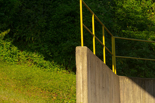 A solid concrete wall featuring a bright yellow railing, complemented by trees positioned in the background of the scene