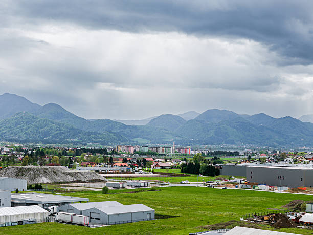 A panoramic view of a green valley surrounded by mountains under a cloudy sky. Industrial buildings and warehouses are visible in the foreground, with residential areas in the distance.