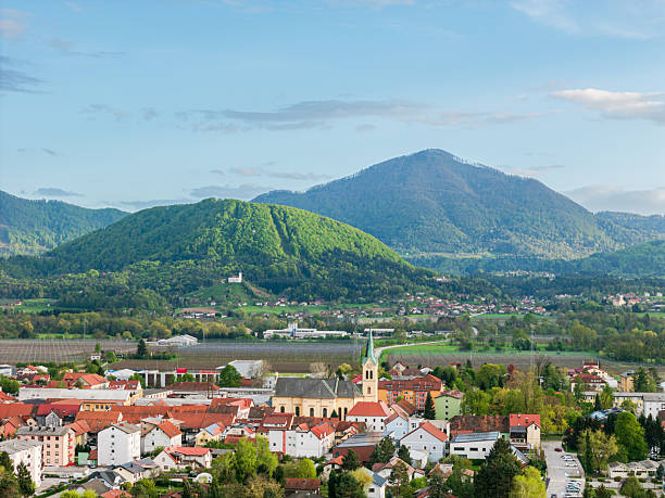 A scenic view of a small town nestled in a valley, surrounded by lush green mountains. The town features traditional architecture with a prominent church steeple. The sky is partly cloudy, suggesting a pleasant day.