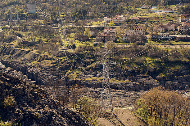 Power lines crossing a picturesque valley, featuring a dam and a quaint village under a bright, sunny sky, create a stunning landscape