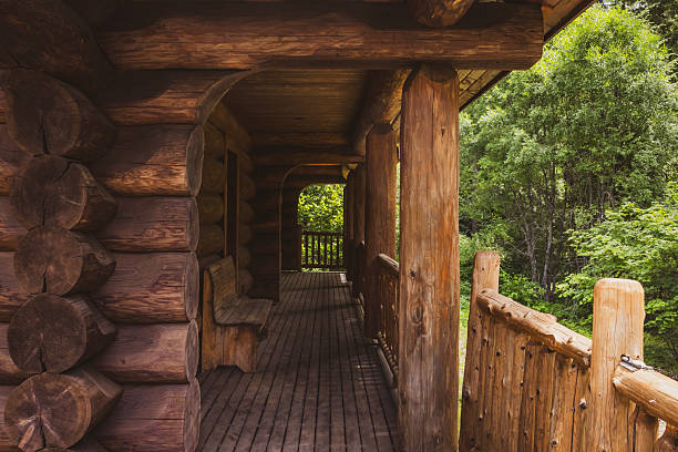 Traditional log cabin with weathered wooden exterior, featuring central door, stacked firewood, and covered porch. Located in open grassland with distant landscape and blue sky background, suggesting isolated rural setting.
