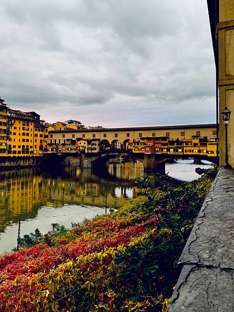Historic architecture lines the Arno River under a vibrant blue sky in Florence, Italy. Painted buildings reflect the charm of Florence's rich cultural heritage, with a scenic stone bridge completing the peaceful riverscape.