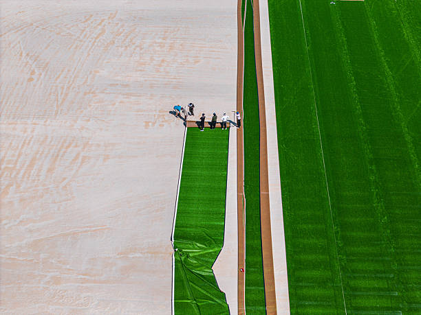 Aerial view of workers laying down artificial turf on a sports field. The contrast between the green turf and the sandy surface is striking, showcasing the installation process.
