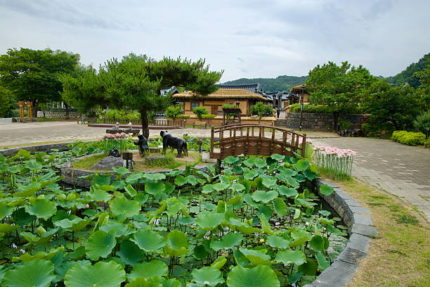 Gongju, South Korea - June 13th 2025: A lotus pond with a wooden bridge and animal statues sits in front of thatched-roof hanok homes and pine trees at Gongju Hanok Village.
