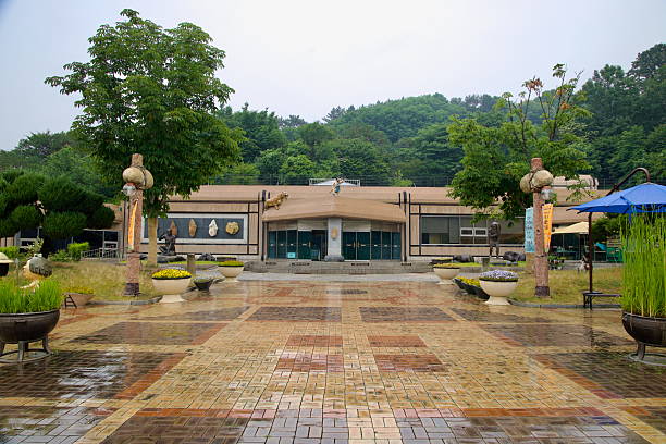 Gongju City, South Korea - June 13th 2025: A wide courtyard leads to the entrance of Seokjangni Museum, lined with flower planters, prehistoric sculptures, and banner poles on a rainy summer day.