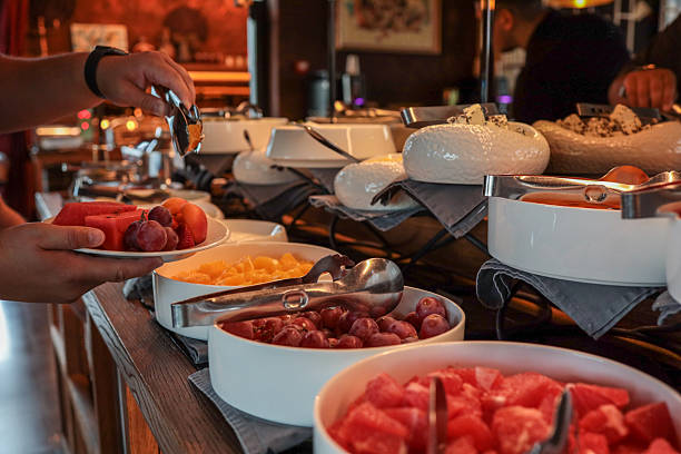 istock Person serving fresh fruit at a breakfast buffet. Plates filled with grapes, watermelon, oranges, apricots, and strawberries. Elegant setting with bowls and tongs on wooden counter 2224573043