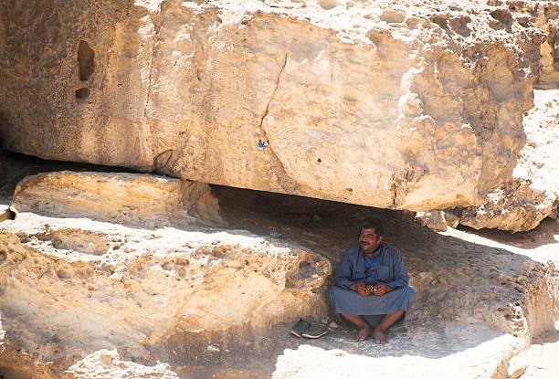 Giza, Egypt - July 05, 2025: Arab man seeking shelter from the heat in the shadow of a huge rock in the Egyptian desert at Giza