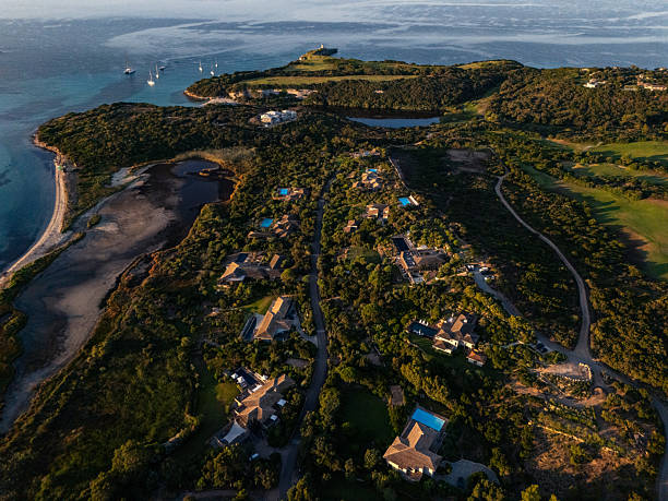 Aerial view of a coastal island featuring private villas with swimming pools surrounded by dense greenery. Scenic ocean waters with anchored sailboats and distant mountain horizon. Tropical vacation destination.