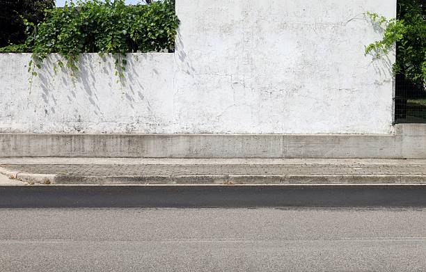 istock Weathered white plaster wall at the roadside with vine on top. Concrete tiled sidewalk and street 2224589668