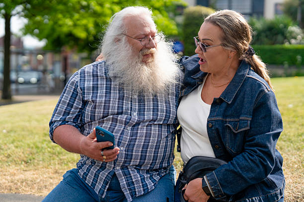 Two individuals are seated in a green park, sharing a joyful moment as they look at a smartphone while basking in the warm sunlight.