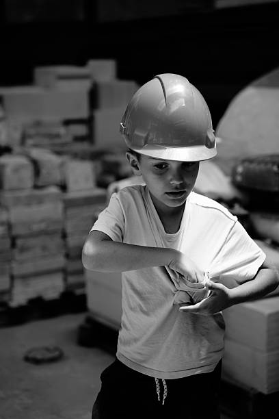 A young boy wearing a construction helmet is searching in his pocket, seemingly looking for a treat. He’s surrounded by piles of concrete blocks.