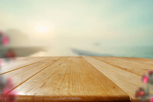 Empty wooden table surface in the foreground with natural textures and warm tones. The softly blurred background features a scenic view of the sea, rocky mountains, and a bright, sunlit sky. The overall composition evokes feelings of peace, travel, and connection with nature. Ideal for product placement in outdoor, wellness, food, skincare, or vacation-themed advertising.