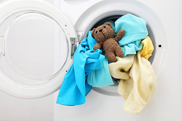 istock Washing machine with clothes and toy bear near light wall, closeup 2224654475