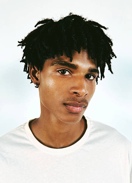 A young man with natural textured hair and warm skin tone wears a simple white t-shirt. He exudes confidence, with a neutral yet engaging expression, set against a clean, light background.
