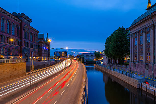 Stockholm, Sweden - June 8, 2025: A scenic view of a city canal at dusk, with light trails from moving vehicles. The sky is a gradient of blue, and historic buildings line the waterway.