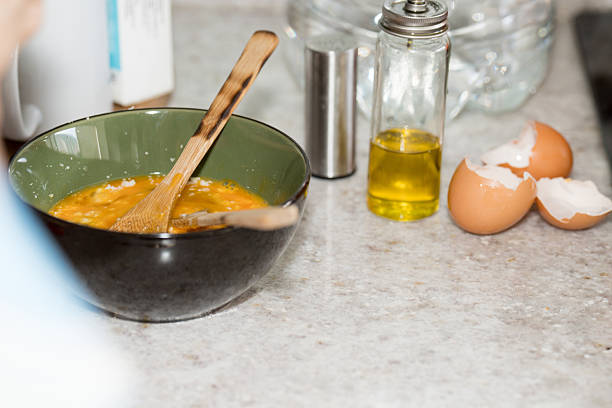 Close-up view of a kitchen countertop showing a green and black bowl with whisked eggs and wooden utensils. Next to it are broken eggshells, a small bottle of olive oil, and a salt shaker. The scene captures a home cooking moment in preparation for a meal, emphasizing fresh ingredients and a cozy domestic environment