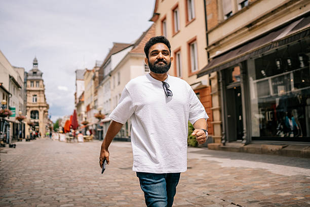 A stylish Indian tourist confidently walks down the cobblestone streets of Landau, Germany, wearing casual attire. The charming urban surroundings reflect a blend of history and modern life.
