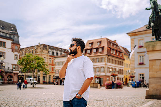 A man stands in the center of a picturesque European square, surrounded by colorful historic buildings and flower-adorned balconies. The bright daylight and clear sky emphasize the charm and architectural heritage of the city.