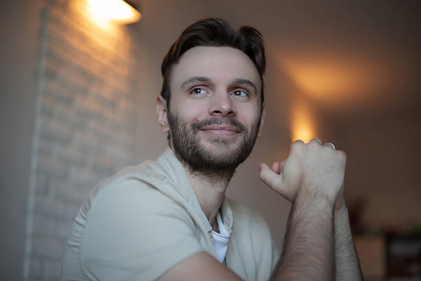 istock Smiling young man sitting indoors in a relaxed pose, looking away with a hopeful expression, enjoying a peaceful moment at home in soft evening lighting. 2224666720