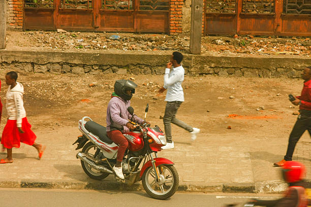 A candid and dynamic street scene captured in Rubavu Town, Rwanda, depicting a lively cross-section of daily life and movement. Centrally, a red motorcycle with a helmeted driver is positioned on the edge of the paved sidewalk. Nearby, two young people walk together, one in a red outfit and another in a white hoodie. On the mid-right, a man in a light blue shirt walks away, while on the far right, another man in a red shirt walks towards the viewer with a blurred motion, indicating the constant flow of activity. The background features a distinctive weathered building constructed from reddish-brown bricks with multiple large, rustic wooden doors, characteristic of the local architecture. The dusty ground and various elements convey the authentic, bustling atmosphere of an urban environment in Rubavu. This image captures the energy, diverse modes of transportation (moto-taxis), and candid interactions of daily life in Rwanda's Western Province. Ideal for themes of urban life, daily routines, African culture, community, youth, transportation, development, street photography, and everyday scenes.
