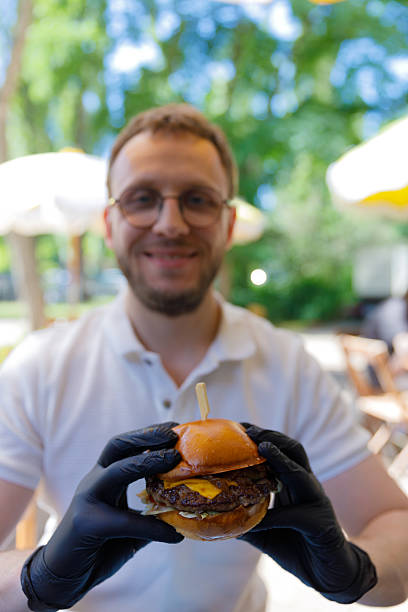 Delicious Cheeseburger Delight: A Focused Enthusiast Grins While Holding a Tempting Burger in a Stunning Outdoor Setting Under Shade and Scenic Background